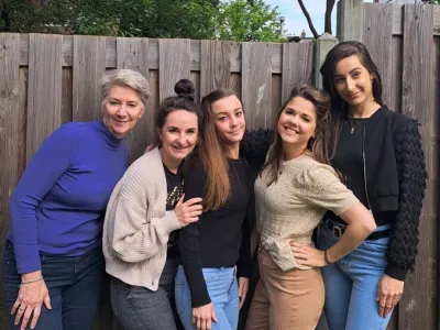 Vijf vrouwen poseren voor een houten schutting met groene bomen op de achtergrond.