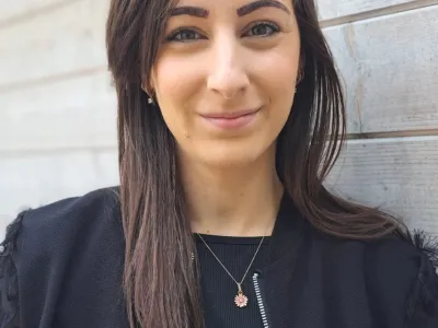 Een portret van een jonge vrouw met lange, bruin haar, die glimlacht en in de camera kijkt, staand voor een houten muur.