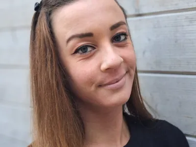 Portret van een jonge vrouw met lang bruin haar, blauwe ogen en een zwarte trui, zittend tegen een houten muur.