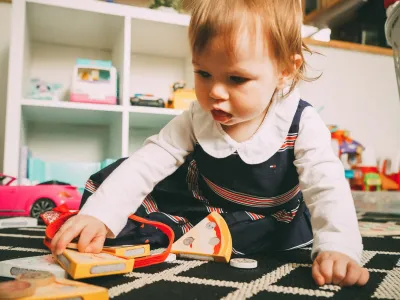 Een peuter die op een zwarte en witte gestreepte tapijt speelt met speelgoed, waaronder een sliert plastic pizza en andere speelgoedstukken, in een speelhoek met witte kasten en speelgoed op de achter