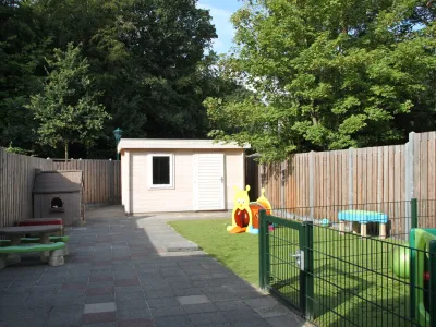 Een kinderopvangtuin met een houten speelhuisje, een kinderzitje en zitbank, een speelgoedhuisje, speeltoestellen en een omheind grasveld, omgeven door een houten schutting en grote bomen.