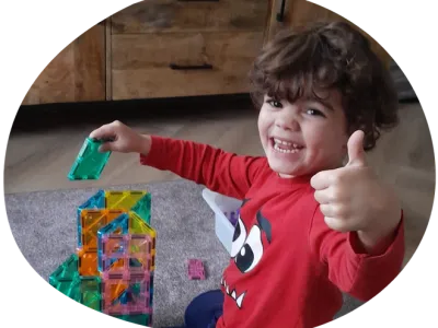 Drie lachende kinderen liggen op een oppervlak, één met een potlood in de hand, omringd door kleurrijke voorwerpen.