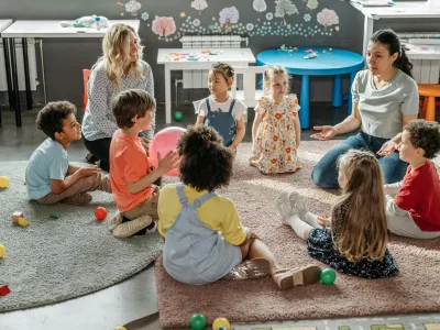 Twee leerkrachten leiden een groep kinderen in een kring op een kleed. Klaslokaalopstelling.