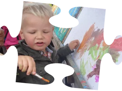 Een jong kind in een roze jasje zit in een rood wagentje, getrokken door een jongen op een gele speelgoedtractor.
