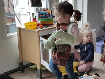 Een kind in een spijkerbroek en een vestje houdt ovenwanten vast en staat bij een speelkeukentje.
