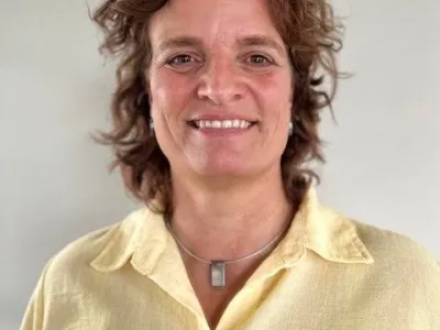 Vrouw met krullend bruin haar, gekleed in een geel shirt, glimlachend naar de camera.