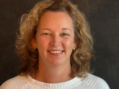 Vrouw met krullend bruin haar, gekleed in een geel shirt, glimlachend naar de camera.
