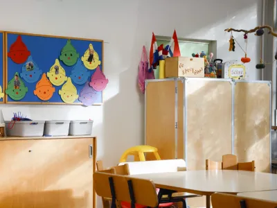 In een door de zon verlichtte ruimte staan tafels en stoelen. Aan de muur hangen kleurrijke voorwerpen en tekeningen.