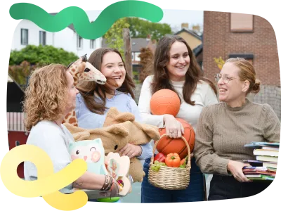 Vier medewerkers van het team van Thornerkwartier staan lachend met de handen vol met spullen voor de kinderen, zoals boeken, ballen, speelgoed, dekens en fruit op het plein voor de school.