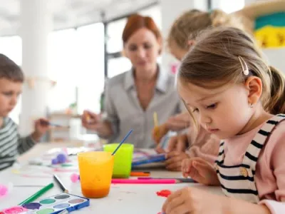 A group of small nursery school children with teacher indoors in classroom, painting.